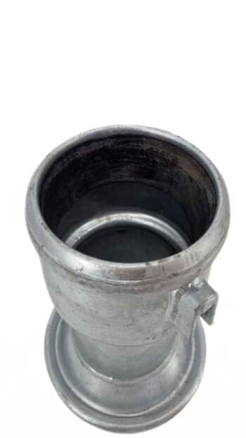 Ein Übergang HK89 Becher auf Hakenverschluss (Schlitz): ein Metallzylinder mit breiter Öffnung, dicken Wänden und einem kleinen rechteckigen Schlitz auf einer Seite, dargestellt vor einem einfachen weißen Hintergrund.