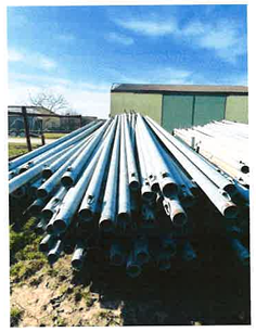 Ein großer Stapel Valducci Alu Rohreinheit DM80, DM100 Metallrohre liegt im Freien auf dem Boden in der Nähe eines grünen Industriegebäudes unter blauem Himmel gestapelt.