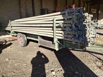 Ein mit langen Metallrohren beladener Anhänger ist in einem Haus auf einem felsigen Boden abgestellt; das Sonnenlicht wirft den Schatten einer Person, die das Foto macht.