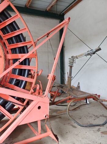 Der Rainstar Typ 82-360 S, ein großer roter Bewässerungsschlauchaufroller mit schwarzem Schlauch, ist an einer Metallregneranlage in einem Lagerraum mit Betonwänden und Metalldach installiert.