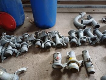 Eine Vielzahl an Beregnungsteilen ist auf einen Betonboden neben zwei blauen Fässern und einem roten Deckel ausgelegt.