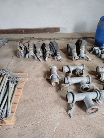 Mehrere Teile der Vielfalt an Beregnungsteilen, darunter Metallfittings und Düsen, liegen auf einem Betonboden; einige Rohre befinden sich auf einer Holzpalette, im Hintergrund steht ein blaues Fass.