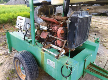 Der COGEM Aggregat 80 PS, ein grüner Generator auf Rädern mit einem Bedienfeld, einem freiliegenden Motor und einem Kühler, steht im Freien auf Erde und Gras.